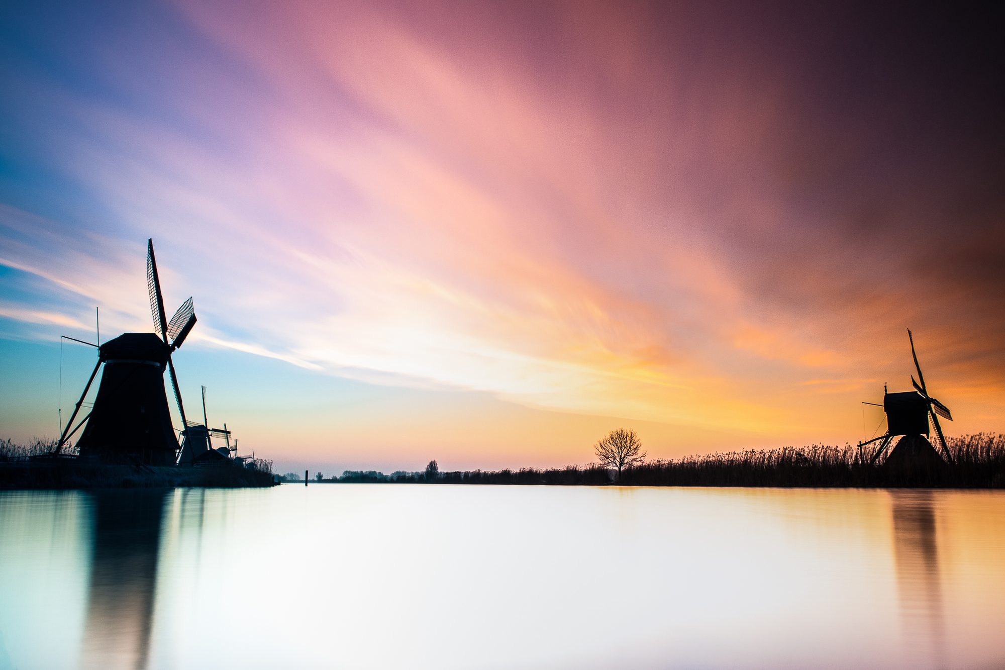 Twee molens in silhouet aan weerszijden van stil water, bij zonsopkomst in roze en oranje licht